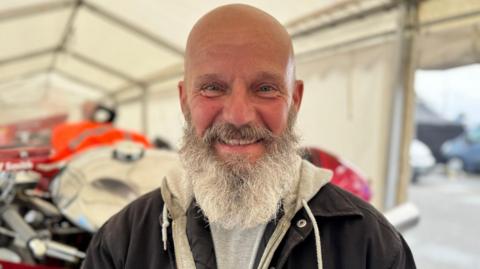 Jeff Smith has a bald head and a lengthy grey beard. He's wearing a dark jacket and standing in a white tent with motorbikes behind him.