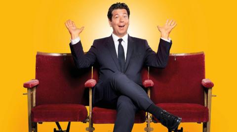 Michael McIntyre sitting cross-legged on a red chair with his hands in the air, posing for a publicity photo. The background is soft yellow. Two more red chairs are placed on either side of him. He is wearing a dark suit.