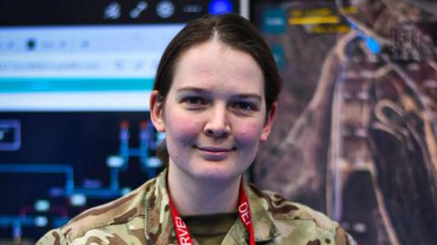 A woman with brown hair and rosy cheeks smiles in an army uniform.