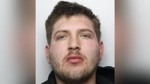 A headshot of a young man with short brown hair, a short beard and moustache and grey eyes.