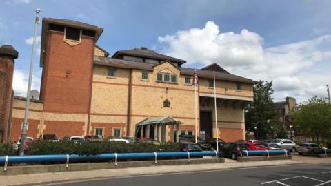 A view of Bedford Prison from outside. It is a a tall brick building with a red brick tower attached to one side. There are cars parked in front of it.