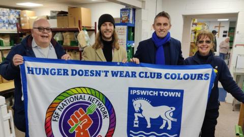 Ipswich Town Fans Supporting Foodbanks stand behind a large flag that they are holding. Ipswich Town manager Kieran McKenna stands with them holding the flag. They all wear winter coats and clothing. The white flag reads: Hunger doesn't wear club colours. It has the Ipswich Town logo on it.