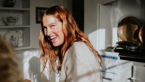 A young woman stands in a kitchen looking at her friend and is laughing. The sun is shining through windows.