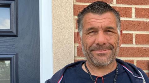 Paul Smith is standing smiling at the camera. He is wearing a t-shirt with a picture of palm trees on it and a navy hooded jacket. There are keys and earphones around his neck. Behind him is part of a black door with windows and a brick wall with a sign on that reads 266D Eastfield Road. 
