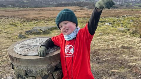 Arthur, eight, stands on an elevated hill area next to a stone sculpture, wearing a woolly hat, red t-shirt and gloves, with his hand in the air. 