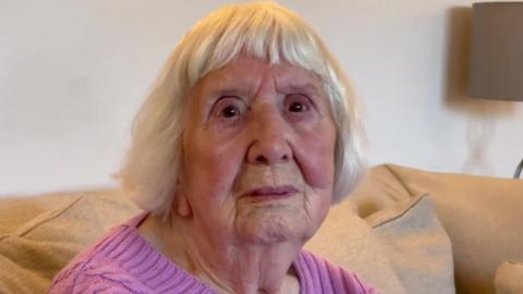 Betty Brown with short cropped hair and a pink jumper, sat on a sofa