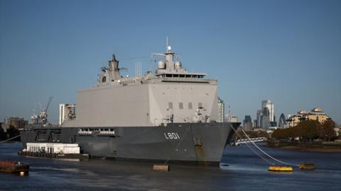 A large naval vessel, anchored in the River Thames with a city skyline in the background, under a clear blue sky.