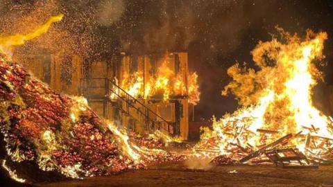 An industrial site, including a portacabin and large piles of wood and woodchip, fully alight. It is dark, with large orange flames stretching into the sky.
