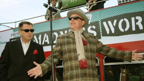 Two men stand in front of a red double-decker bus with a sign reading ‘MADNESS WORLD’. The man on the left is wearing a black suit with a red pocket square, a white shirt and black sunglasses. The man on the right wears a plaid jacket, a scarf with red fringe details, black sunglasses, and a grey hat. A microphone stand is in front of them, and musicians are visible on the upper deck of the bus playing instruments.