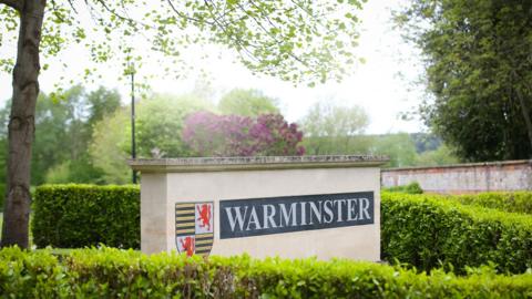 A sign on a thick brick rectangular object which says 'Warminster' with a hedge and trees in the background.