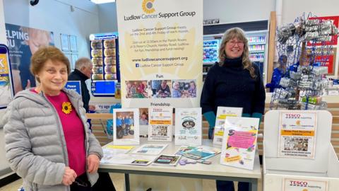 Support group member Sybil Marsh and artist Miranda Goudge stand next to a recycling awareness stand at Ludlow Tesco. There are leaflets on the table and there is a display stand behind them. The shop can be seen in the background.