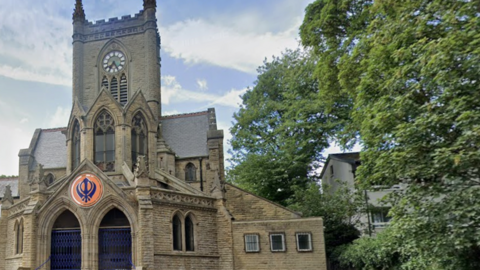 A brown stone church building with a clock tower, and a large orange and blue Sikh symbol over the front door.