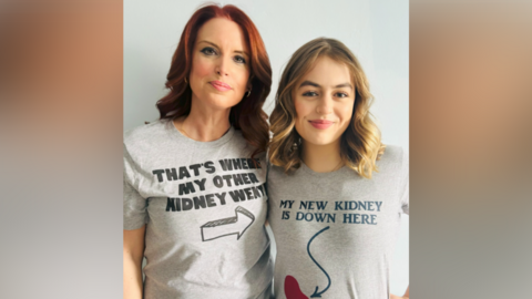 Emma Palmer is standing side-by-side indoors with Maya. Emma has red hair and is wearing a grey T-shirt with writing printed, that reads THAT'S WHERE MY OTHER KIDNEY WENT. It is accompanied with the drawing of an arrow, pointing to her left. On her left is Maya, who has blonde hair and is smiling. She has a grey T-shirt reading MY NEW KIDNEY IS DOWN HERE, with an arrow pointing to a drawing of a kidney, which is coloured in red.