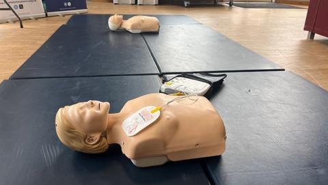 Two female CPR mannequins lie on a blue mat in a classroom with a wooden floor. A mobile defibrillator is attached to the torso nearest the viewer