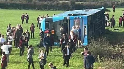 A blue double-decker bus overturned overturned in a field, multiple pupils are gathered around the bus looking at the crash.