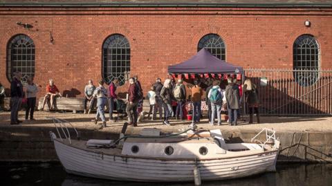 A group of people crowded around a stall with bunting on its edges. A white boat is in the water in the foreground.