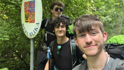 Kyle Spring (L), James McCarthy-Hill, and Adam Mitchell (R) wearing large back packs and posing for a selfie beside the Czech Republic border sign in front of lots of trees.