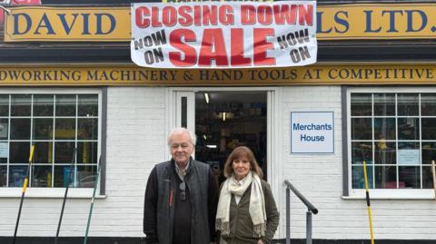 (l-r) David and Maureen Hunt looking at the camera, with the tool shop behind them.