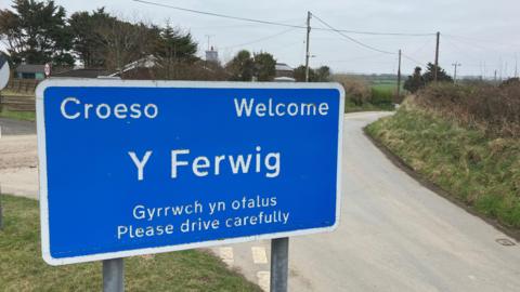 A blue road sign which says Y Ferwig, the name of the village with language in English and Welsh which says 'welcome' and 'please drive carefully' 