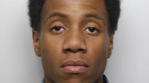 Romaine James' mugshot, where he is looking at the camera with a serious expression in front of a grey background. He has black hair.