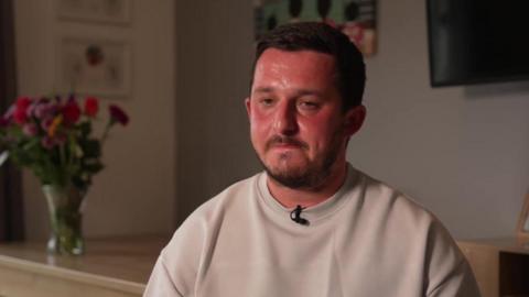 a man sitting in a darkly lit room. He is wearing a beige sweatshirt. His face is reddened. A wooden table is behind him. A glass vase of flowers is on the table and there are pictures hanging on the wall.