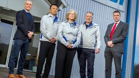 Executives are standing outside the Kasai building. One man on the far left of the picture is in a suit jacket and jeans, the three in the middle are all wearing matching waterproofs and the man on the right is in a full grey suit.