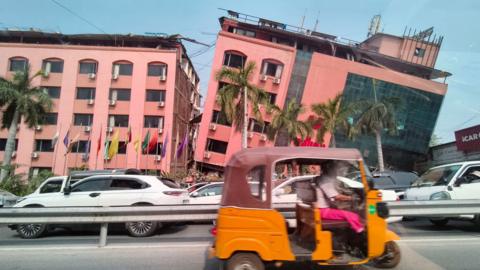 Two pink large buildings next to each other. One is at a slant and looks like it has been toppled onto its side. A road is in front of the buildings.