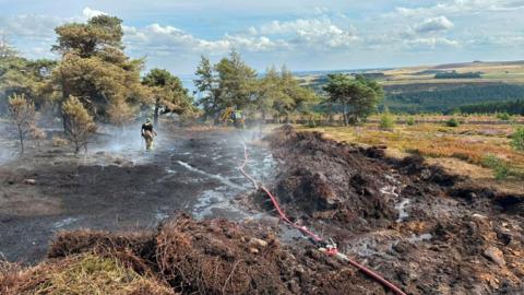 A moorland field has been half burned away with just blackened soil. In the distance is a firefighter and a tractor by some trees