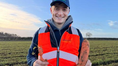 A man with a blue coat and blue cap is standing in a field holding a large carrot in one hand and a deformed carrot in another. He also has an orange hi-vis vest on. He is smiling and looking into the camera.