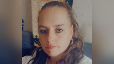 Ms Blackham is smiling in a selfie photo. She has brown hair tied back and is wearing a white top and is sat inside a home.