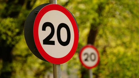 Close up of a round 20mph road sign with blurred greenery behind