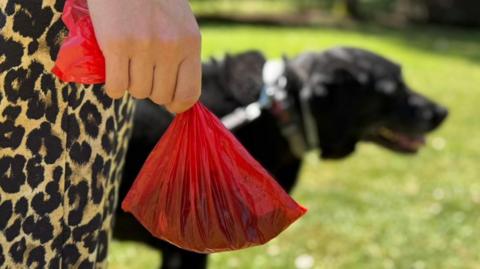 A close-up of a woman's hand holding a red poo bag. She wears leopard print trousers. A dog can be seen in the background, standing in a grassy field.