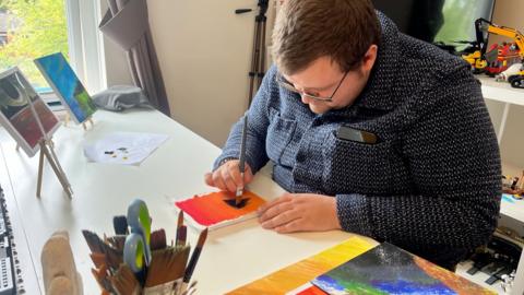 a man sat at a table painting. He had a dark top on, glasses and brown hair.