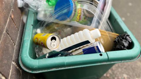 Mixed recycling including a drinks can, shampoo bottle, plastics, jars and a small piece of waste covered in a black bag on the pavement in a green box