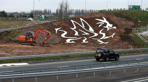 The eastbound side of the A494 dual-carriageway in Flintshire showing work being completed on a white dragon shape cut into a sloping embankment. The design is filled with white stone so that it stands out to passing motorists. A digger can be seen on the muddy ground.
