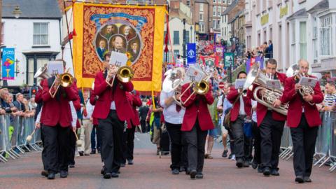 The Durham Miners' Gala. A brass band is walking down the street in red blazers. A red, yellow and blue miners' banner with five men depicted in the middle is being held up behind them. More banners and people can be seen walking behind them with people behind barriers on either side of the street.