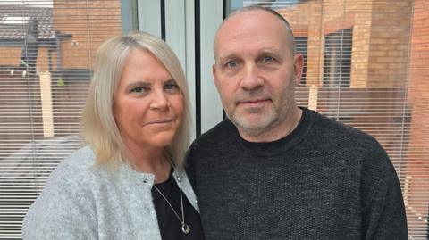 Wendy Leech, 55, who has blonde hair and wears a light grey cardigan, and Paul Leech, 60, who has short grey hair and wears a dark grey jumper, stand side-by-side in front of a set of patio doors looking out into a garden.