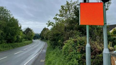 A square digital red flag attached to a tall pole next to a road, which is lined by greenery and trees. 