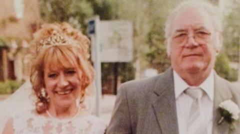 Karen Teasdale-Robson in a white dress and veil smiling next to her father Brian Robson, an older man with white hair and glasses wearing a suit, tie and a carnation button hole.