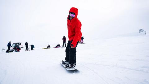 A boarder in a red jacket and black trousers on a board in snow at Cairngorm Mountain.