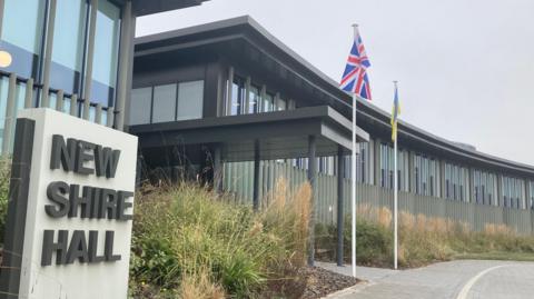 A sign saying 'New Shire Hall' is to the left with a green and grey building to the right. A variety of foliage is in front. There are two flag poles, one with the union flag and the other with the blue and yellow colours of the Ukrainian flag. 