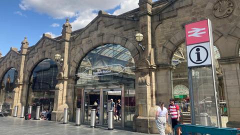 The outside of Sheffield railway station with large glass windows and stone arches. People are milling around