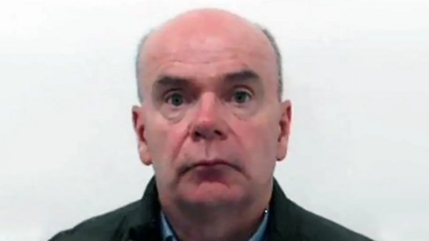 a police mugshot of Daniel Doherty. He has a bald head and is wearing a green jacket over a blue shirt