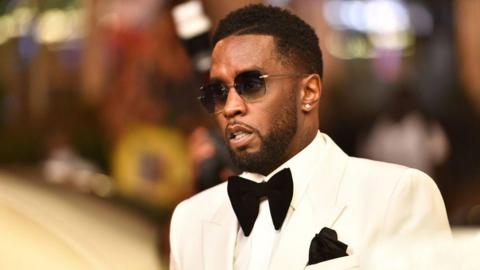 Sean "Diddy" Combs photographed in 2021. He wears sunglasses, a white tuxedo with a large black bow tie and matching pocket handkerchief. 