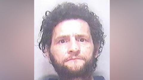 A police mugshot of a man with untidy black hair and a short beard. He is staring directly into the camera.