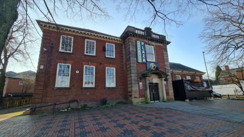 A two-storey brick building of imposing early 20th Century civic style, sat in a small plaza area with more modern residential housing nearby