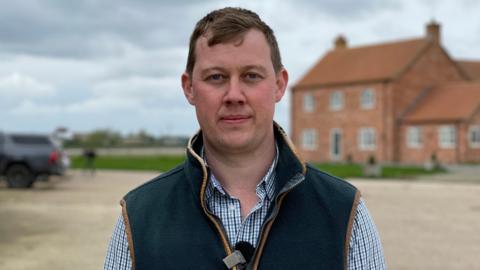 Mr Ward is wearing a check shirt and a dark green gilet, he is standing in front of a red brick farmhouse. In the distance a grey 4x4 car is parked 