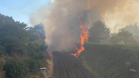 Flames tower above a large hedgerow on the right side of a railway line. A huge plume of smoke completely blocks the view up the line.
