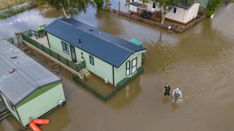 People wade through floodwater surrounding mobile homes at Cogenhoe Mill Holiday Park after the River Nene burst its banks, on September 24, 2024 in Cogenhoe, England