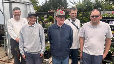 Five men, varying in age, standing in a row in front of stand of plants
